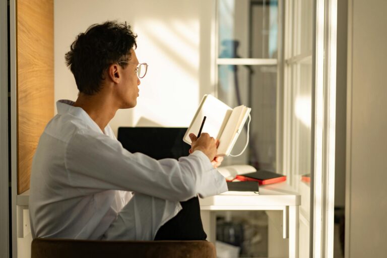 A young man sitting by a window in a sunlit home office, writing in a notebook.