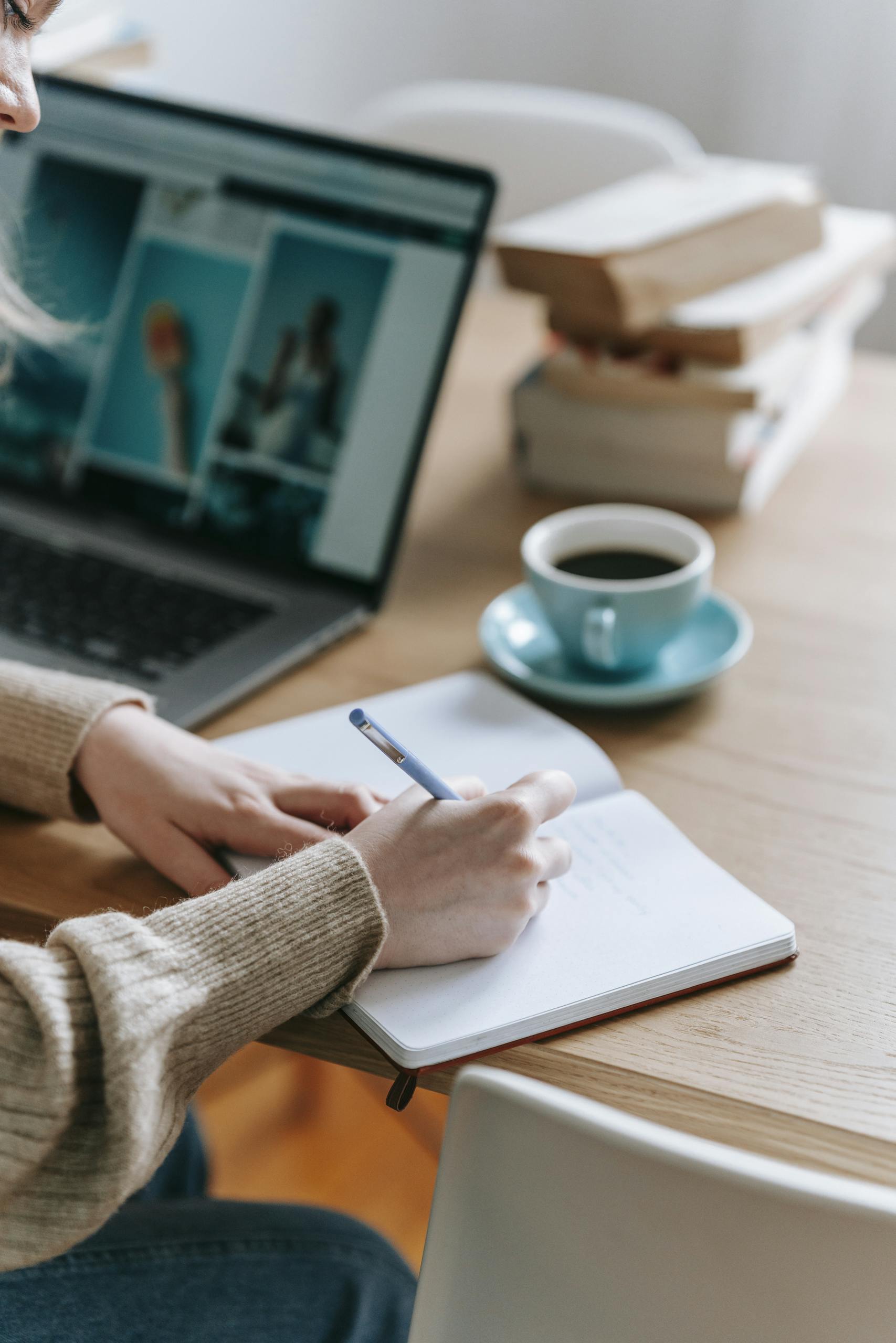 Close-up of a person writing in a notebook beside a laptop and cup of coffee on a desk.