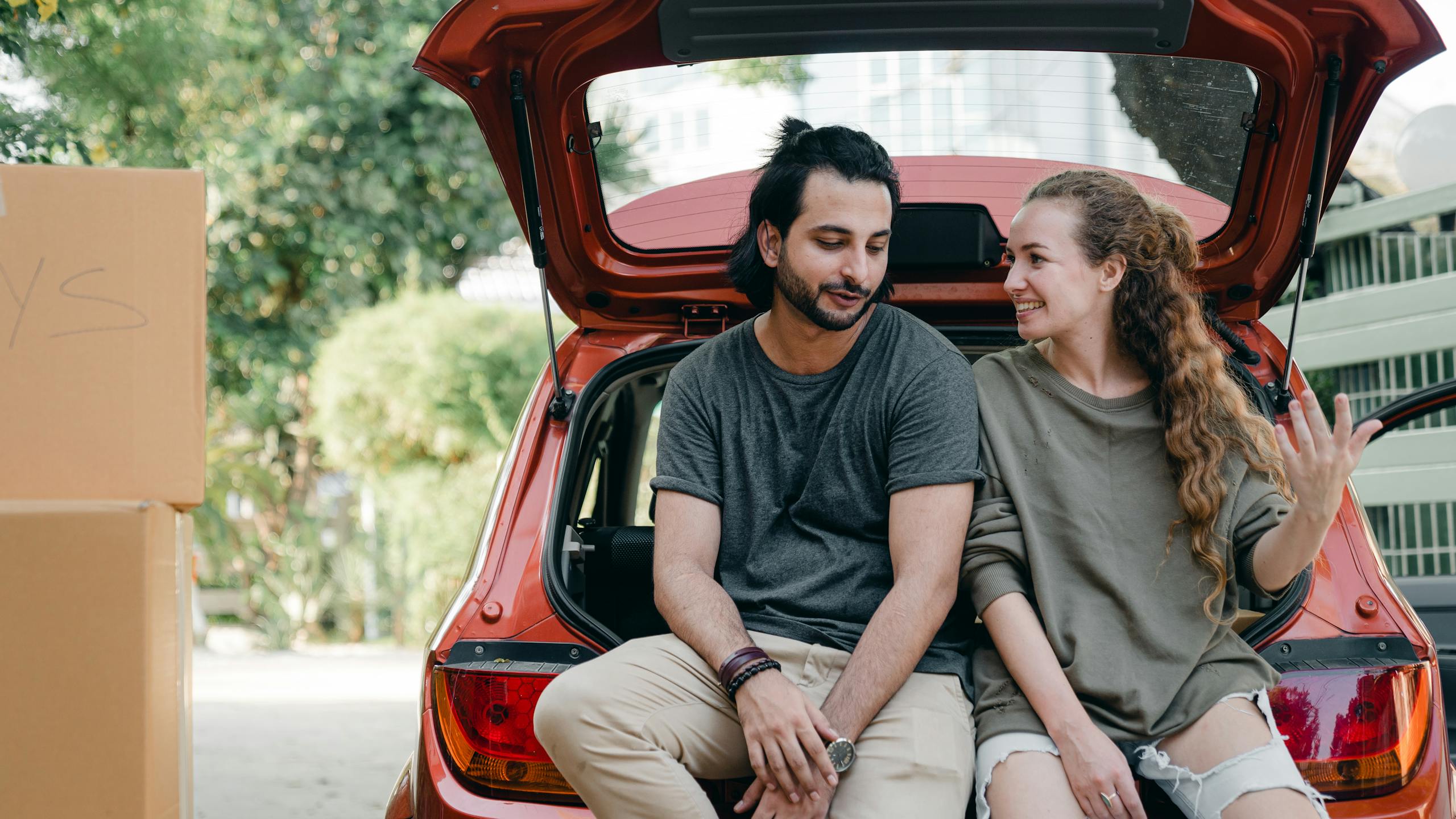 Happy couple sitting on car trunk with boxes, enjoying move-in day together.